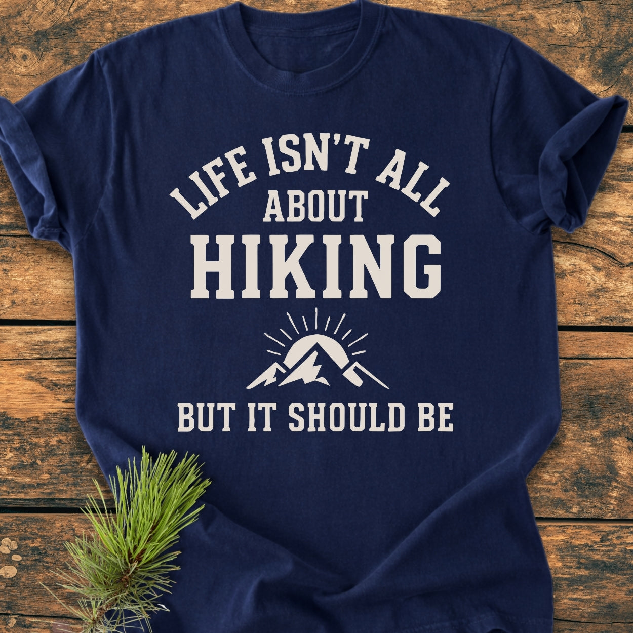 Life Isn't All About Hiking - Vintage Wash Tee on a wooden surface, features a white graphic with mountains and sun rays. Pine sprig accents the soft, relaxed-fit, navy-blue T-shirt.