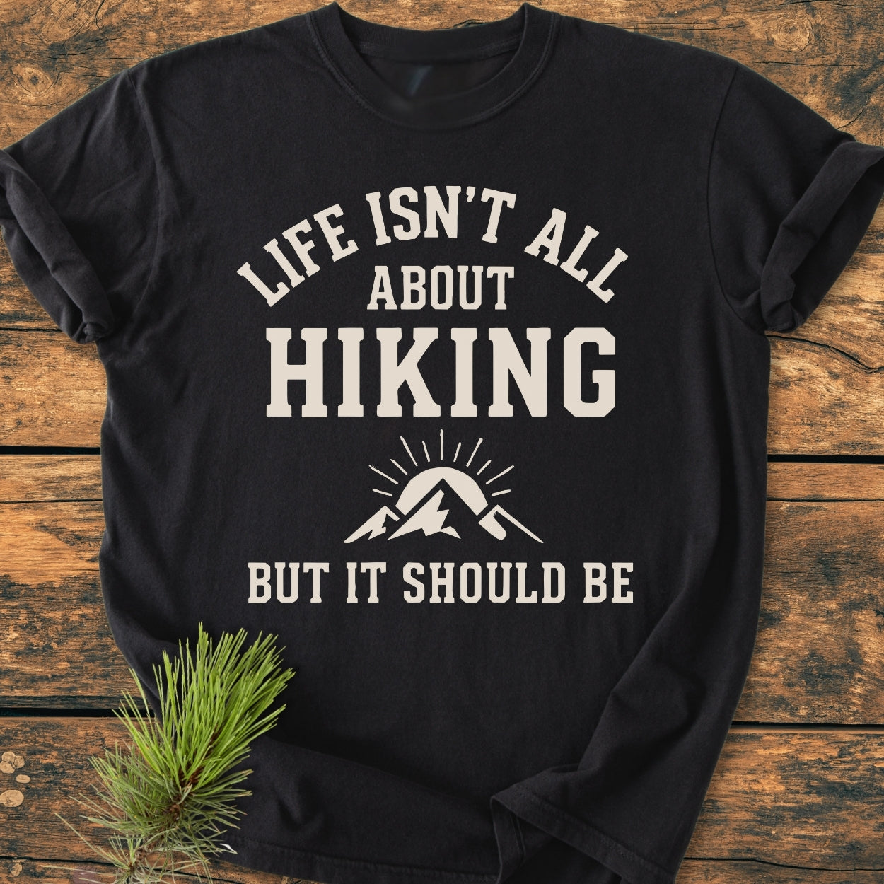 Life Isn't All About Hiking - Vintage Wash Tee with an off-white mountain/sun graphic on black. Laid flat on wood with pine sprig, exuding a rustic, outdoor vibe.