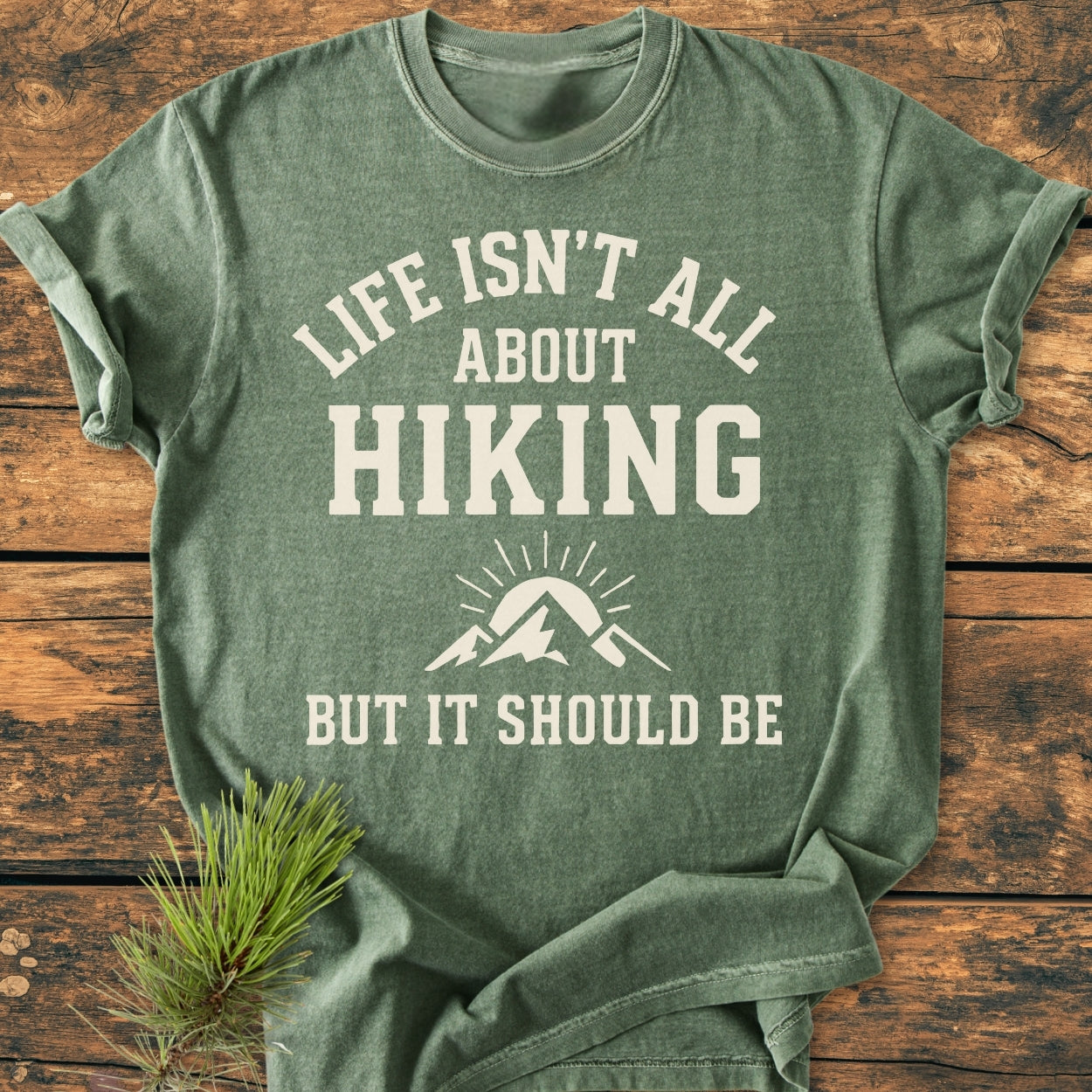 Life Isn't All About Hiking - Vintage Wash Tee featuring off-white text and a mountain-sun graphic on a soft-green, crew-neck design, laid flat on a wooden surface for display.