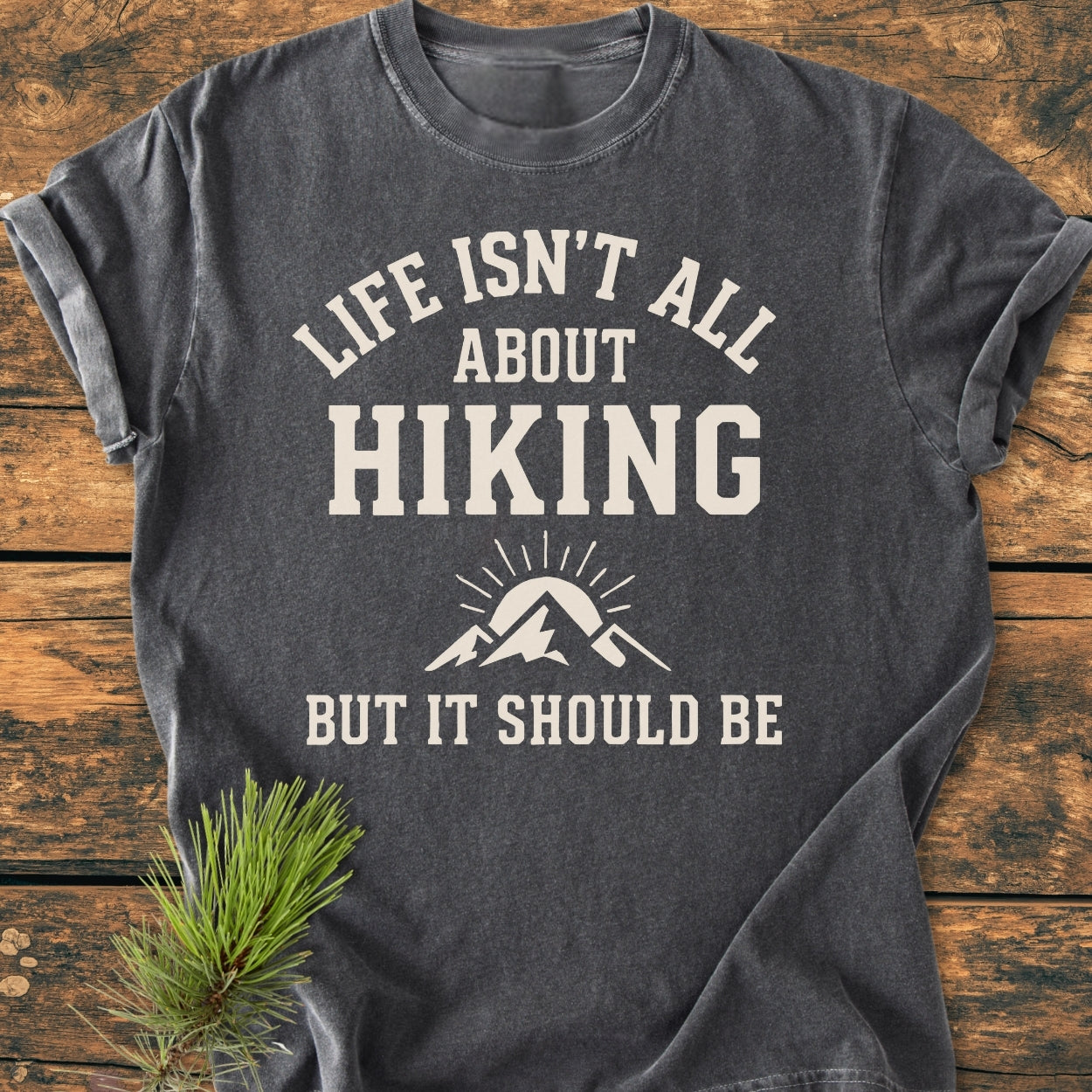 Life Isn't All About Hiking - Vintage Wash Tee displayed on wood, features off-white slogan and mountain-sun graphic. Accompanied by a pine sprig, highlighting the tee's relaxed, soft-washed design.