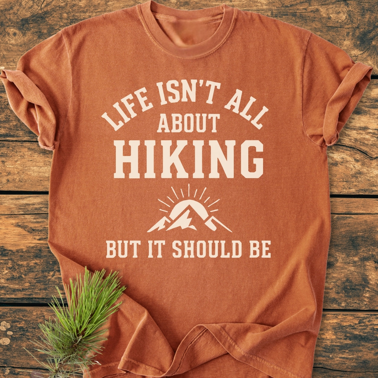 Life Isn't All About Hiking - Vintage Wash Tee, featuring white slogan and mountain graphic, folded on wooden surface with pine sprig. Made from soft 100% ring-spun cotton, relaxed fit for comfort.