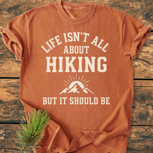 Life Isn't All About Hiking - Vintage Wash Tee, featuring white slogan and mountain graphic, folded on wooden surface with pine sprig. Made from soft 100% ring-spun cotton, relaxed fit for comfort.