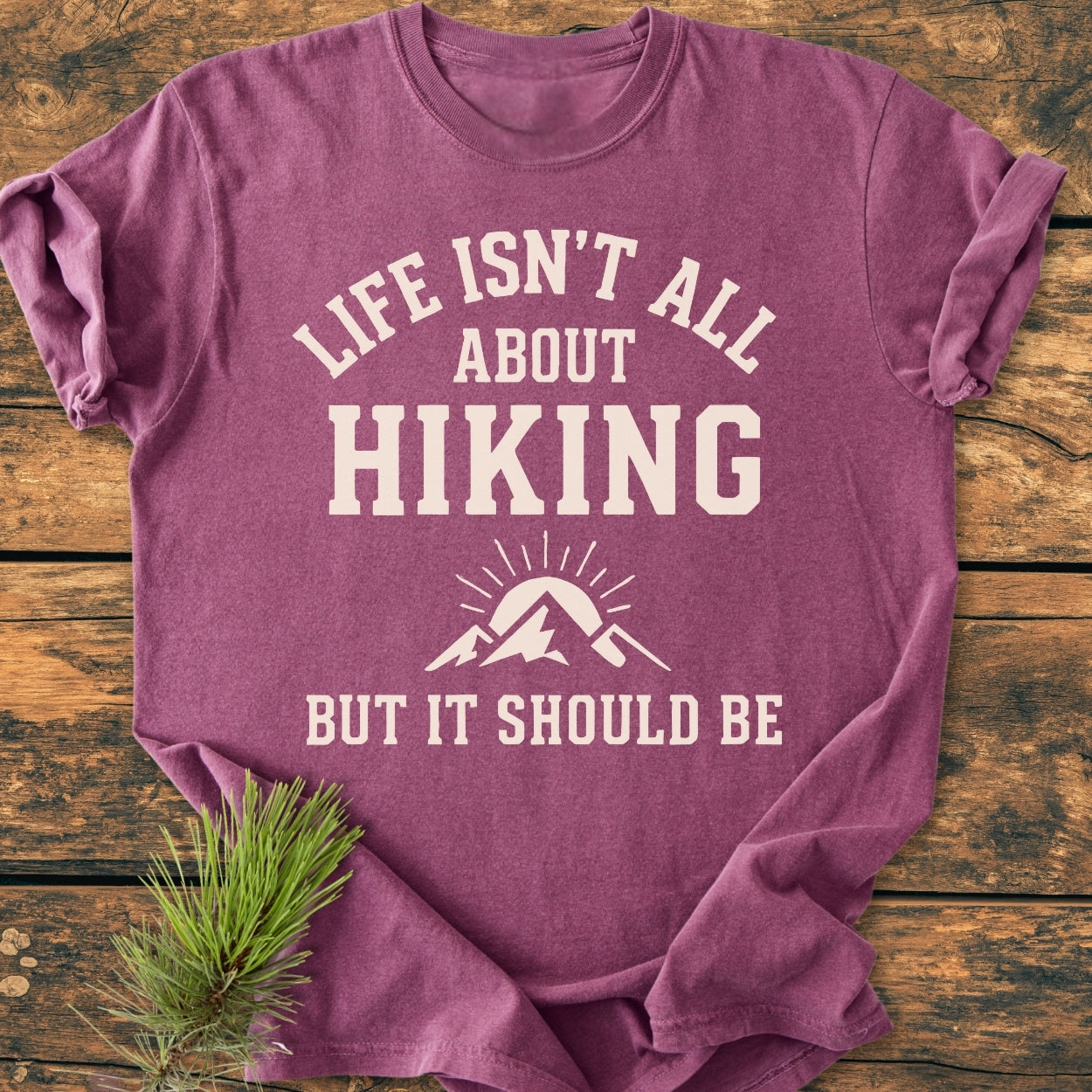 Life Isn't All About Hiking - Vintage Wash Tee, heathered purple, laid flat on wood with pine sprigs. Features bold white slogan and mountain graphic, highlighting its relaxed fit and durable construction.