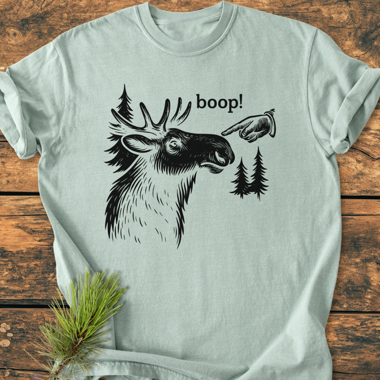 BOOP Moose Funny - Vintage Wash Tee featuring a stylized moose head and boop! text printed in black on a light-green T-shirt, laid out on a wooden surface with a pine sprig.