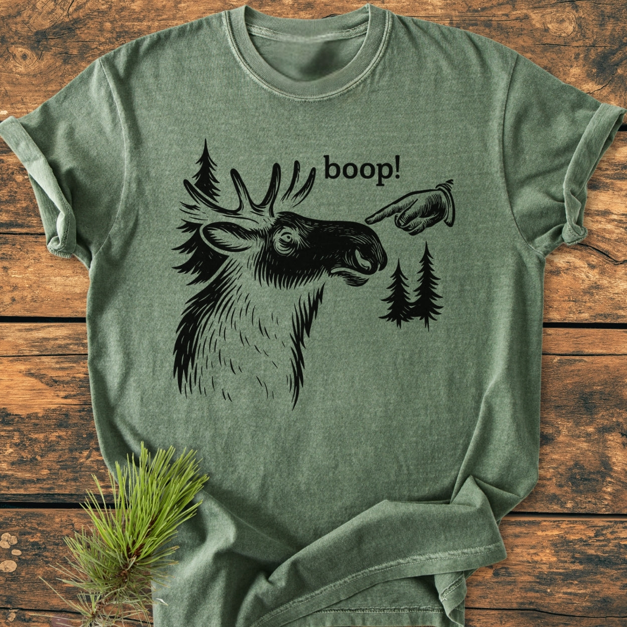 BOOP Moose Funny - Vintage Wash Tee features a stylized moose print with 'boop!' text, laid on wooden background with pine sprig, showcasing its relaxed, durable design in soft-washed, garment-dyed fabric.