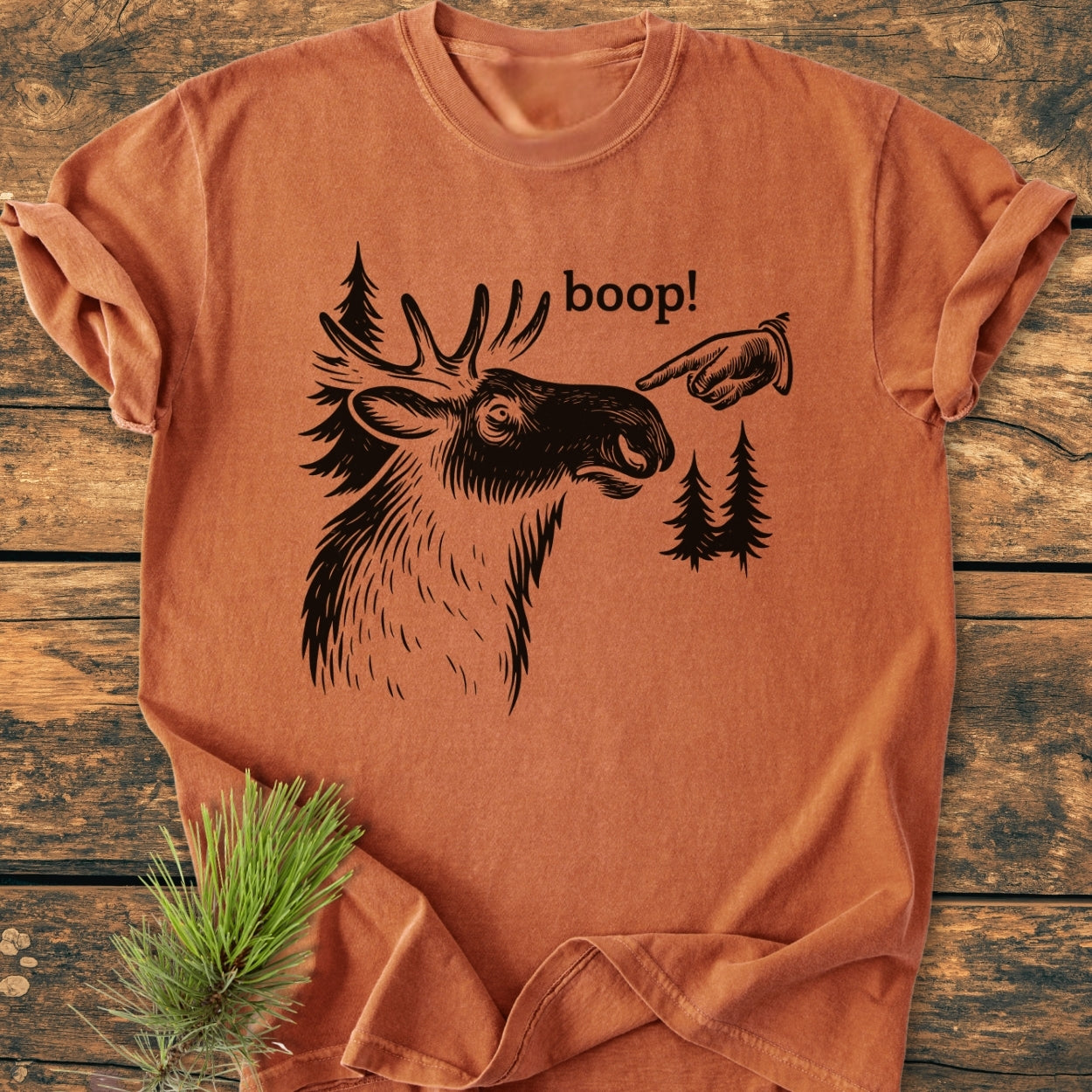 BOOP Moose Funny - Vintage Wash Tee features an orange-rust color with a black screen-printed moose head, pine trees, and boop! on the chest, laid flat on a wooden surface.