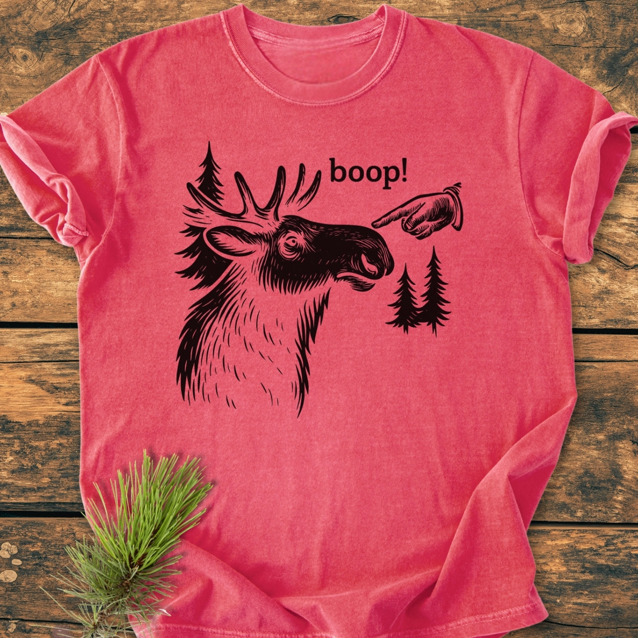 BOOP Moose Funny - Vintage Wash Tee features a moose head line-drawing with a boop! label, printed on a pink-red, short-sleeve, soft-washed cotton shirt, laid on a wooden surface with pine.