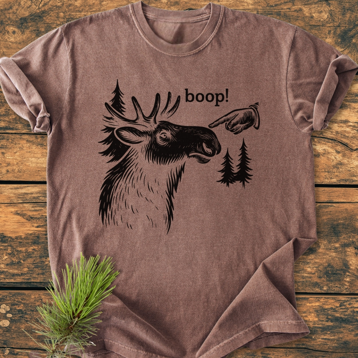 BOOP Moose Funny - Vintage Wash Tee features a stylized moose head, pine trees, and a hand with boop! on a brown, short-sleeve shirt, laid flat with a pine branch accent.