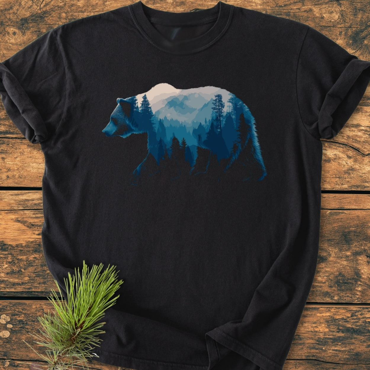 Grizzly Silhouette - Vintage Wash Tee featuring a black T-shirt with a blue mountain-and-forest scene within a walking bear silhouette, laid on a wooden background with a pine sprig accent.