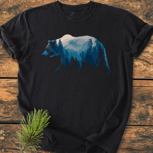 Grizzly Silhouette - Vintage Wash Tee featuring a black T-shirt with a blue mountain-and-forest scene within a walking bear silhouette, laid on a wooden background with a pine sprig accent.