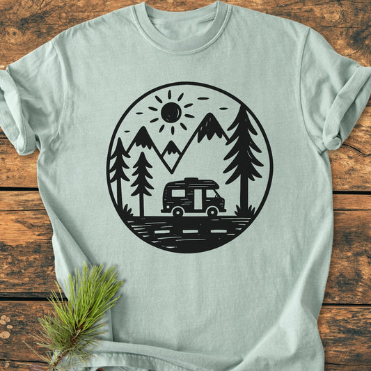 Freedom Is Found Outside - Vintage Wash Tee featuring a minimalist van-life motif with a sun, mountains, pine trees, and a camper van, displayed on a wooden surface with a pine sprig accent.