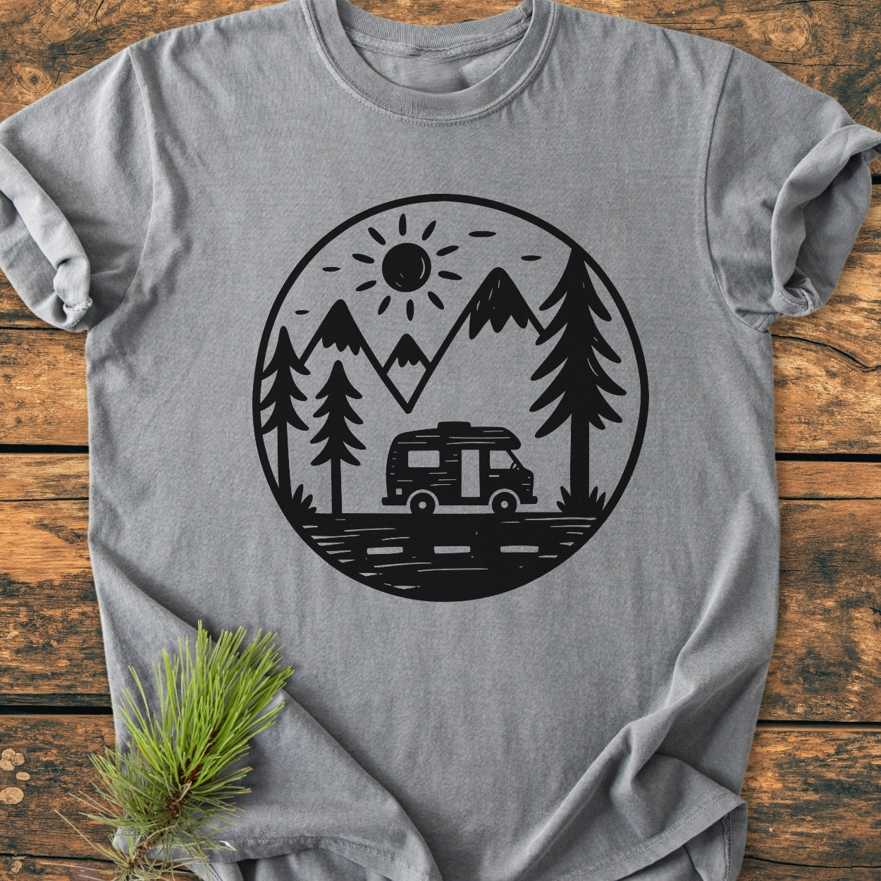 Freedom Is Found Outside - Vintage Wash Tee: A heather-gray T-shirt featuring a black circular outdoor scene with a camper-van, pine trees, mountains, and sun, laid on wood with a pine sprig beside it.