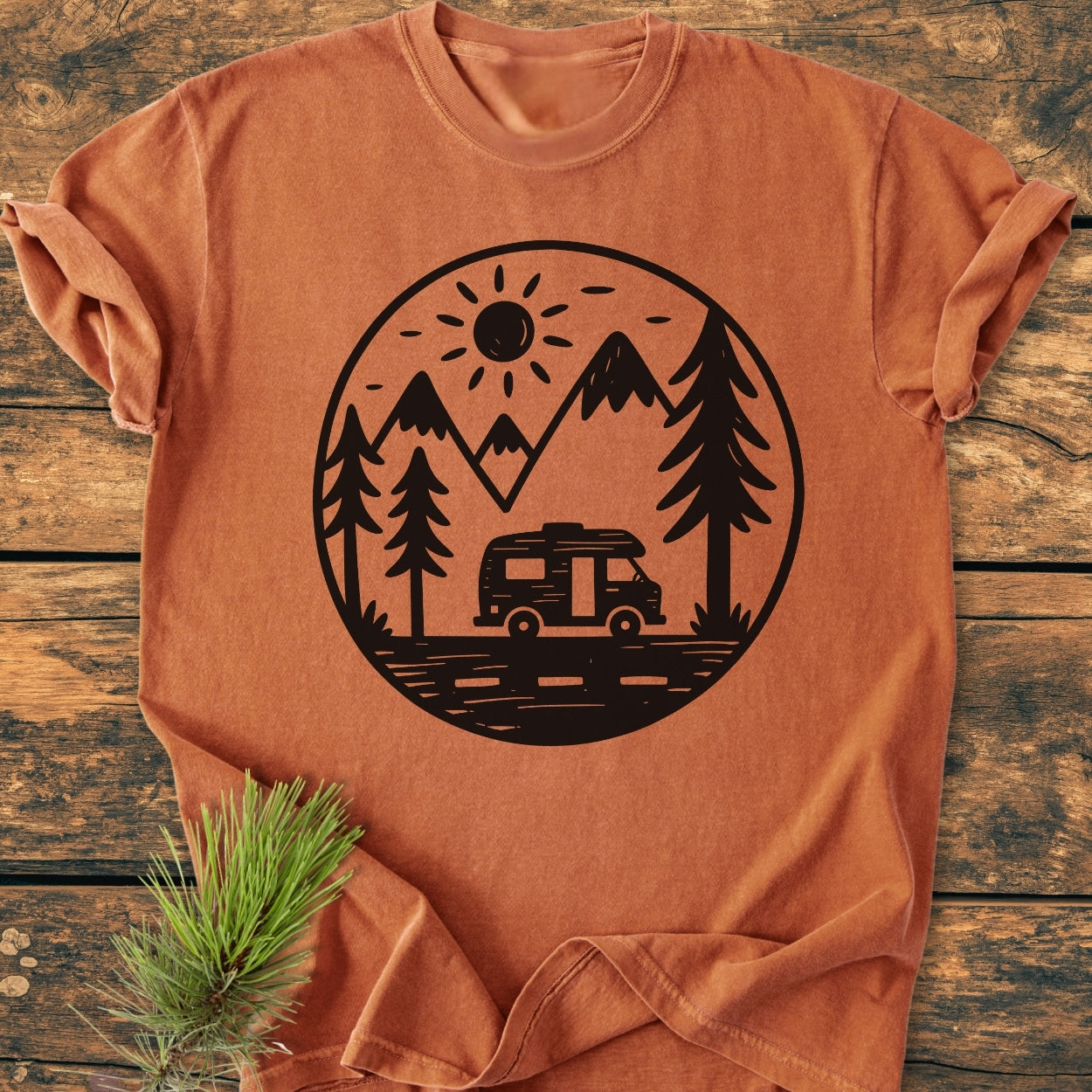 Freedom Is Found Outside - Vintage Wash Tee featuring a black circular graphic with mountains, a sun, pine trees, and an RV, laid on a rustic wood surface with nearby pine sprig.