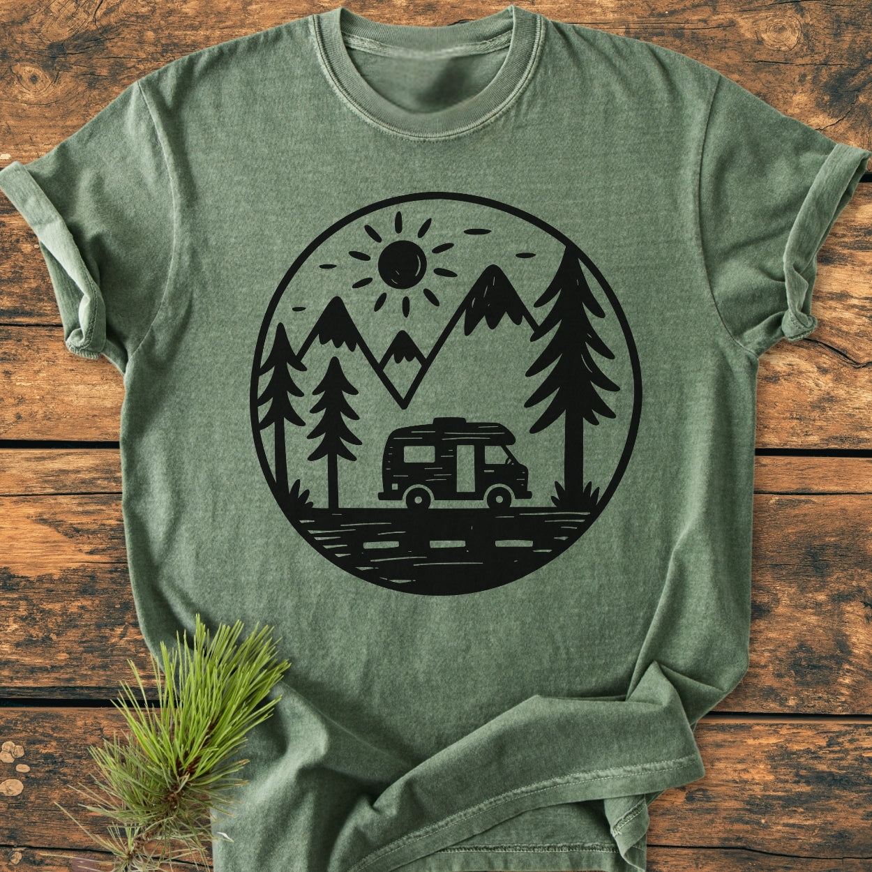 Freedom Is Found Outside - Vintage Wash Tee featuring a black outdoors graphic with a camper van, pine trees, mountains, and sun, displayed on a wooden surface with a pine sprig beside it.