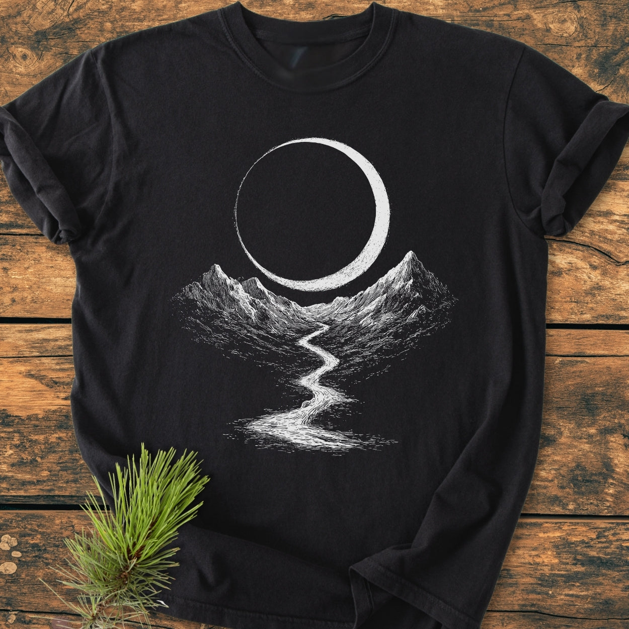 Eclipse Trail - Vintage Wash Tee featuring a white screen-printed landscape with crescent moon, mountains, and river on a black T-shirt, laid on weathered wooden planks with pine needles nearby.