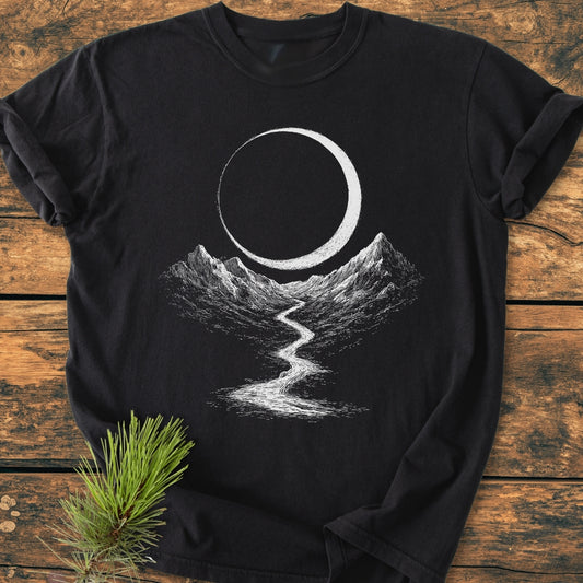 Eclipse Trail - Vintage Wash Tee featuring a white screen-printed landscape with crescent moon, mountains, and river on a black T-shirt, laid on weathered wooden planks with pine needles nearby.