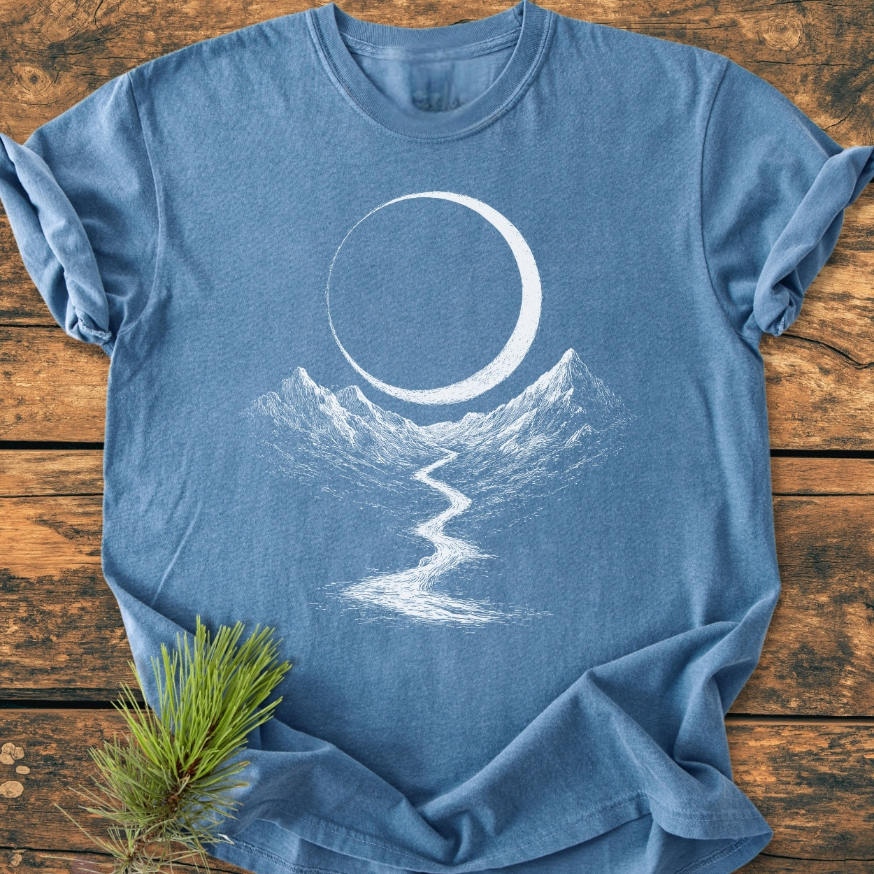 Eclipse Trail - Vintage Wash Tee features a hand-etched mountain scene with peaks, a winding path, and a crescent. Laid flat with a pine sprig accentuating its relaxed, soft-washed design.