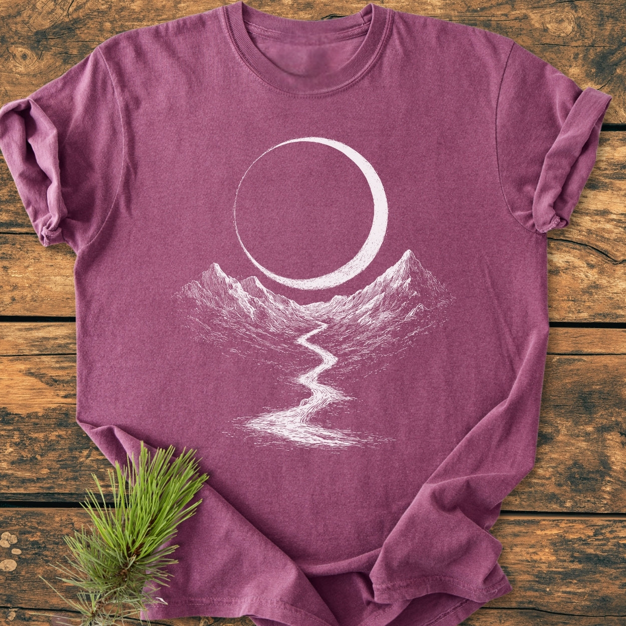 Eclipse Trail - Vintage Wash Tee featuring a crescent moon and mountain design printed on a heathered plum-purple T-shirt, laid on rustic wood, accented by pine needles.