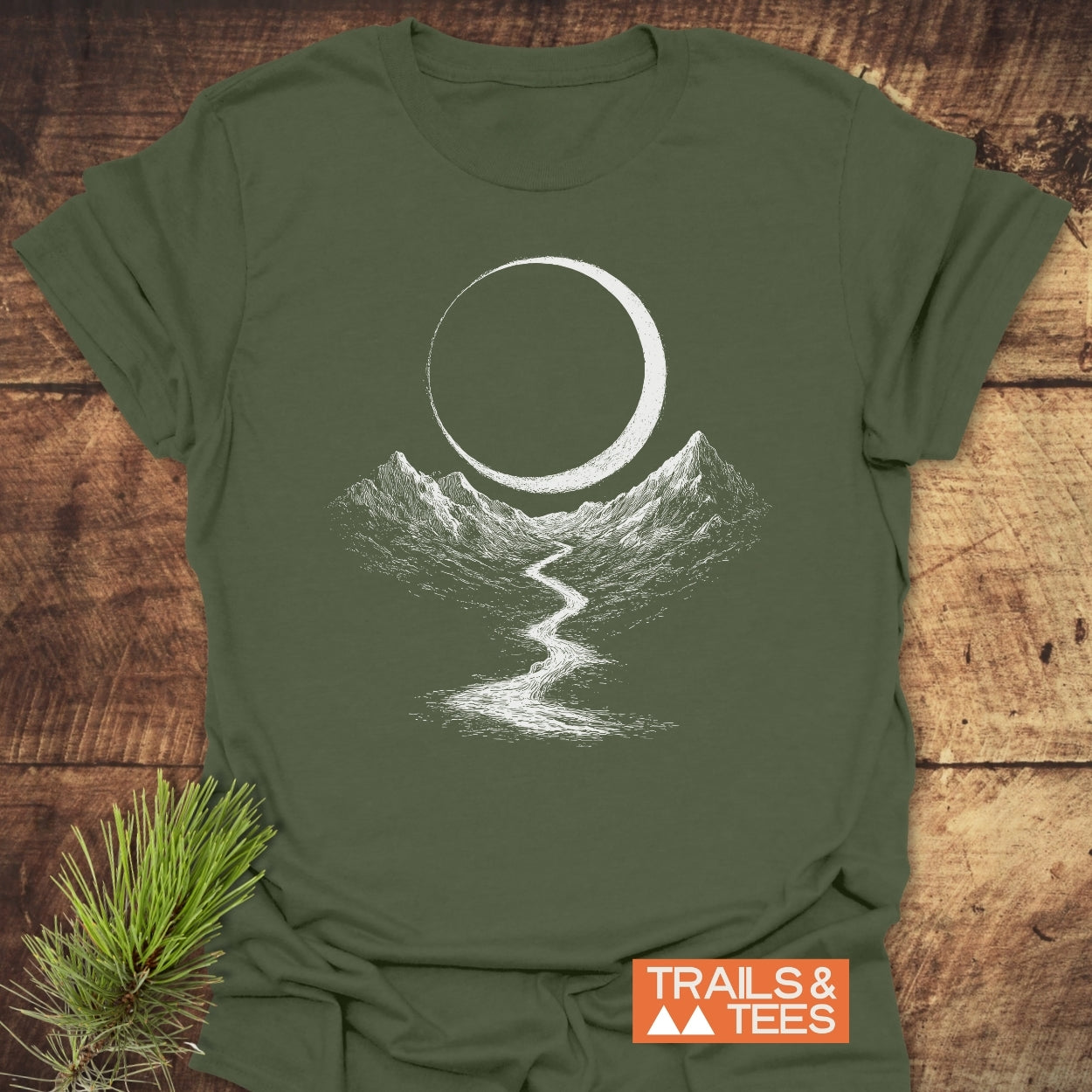 Eclipse Trail T-Shirt on rustic wood, featuring a hand-drawn moon above mountains and a river. Made from soft cotton, perfect for outdoor adventures. Includes TRAILS & TEES logo.