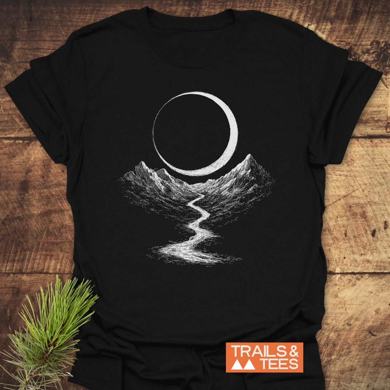 Eclipse Trail T-Shirt featuring hand-drawn mountains, a winding river, and a crescent circle, laid on wood with pine sprig. Crafted from soft, lightweight cotton for outdoor enthusiasts.