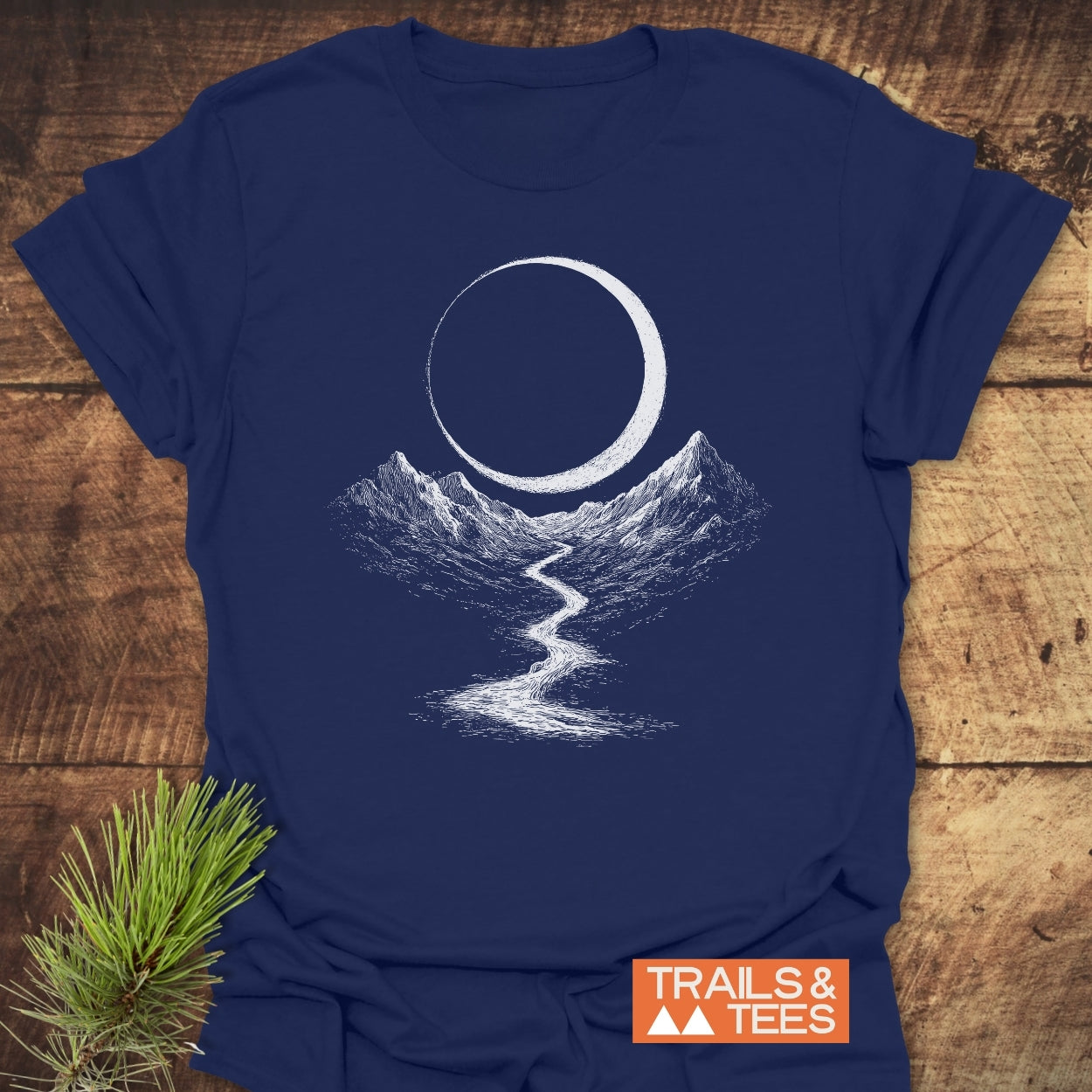 Eclipse Trail T-Shirt featuring a white crescent moon and mountain graphic on navy-blue fabric, laid on rustic wood. Includes a small TRAILS & TEES logo and pine needles beside it.