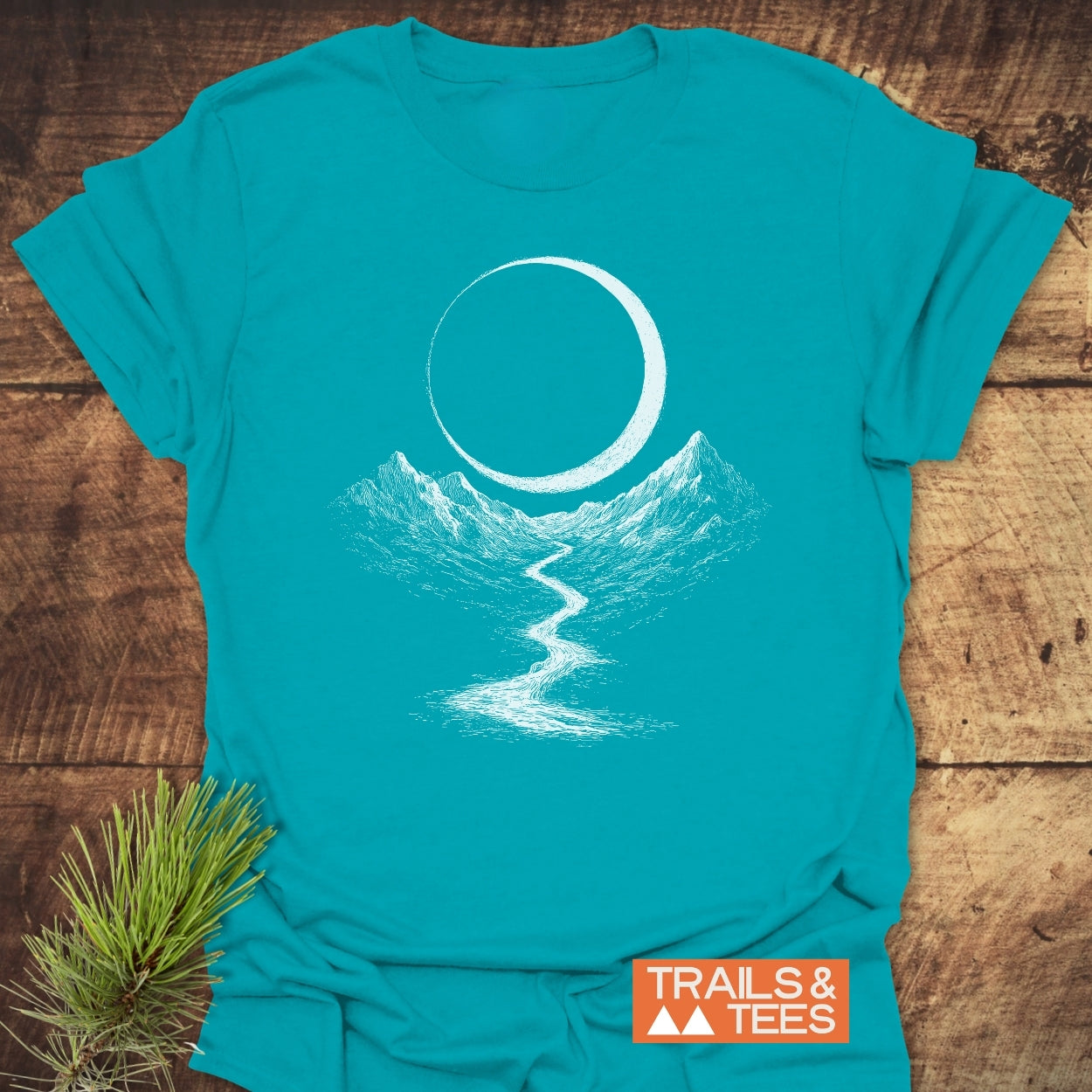 Eclipse Trail T-Shirt featuring a white line-art scene: crescent moon, mountain range, and river. Includes an orange TRAILS & TEES label, crafted for comfort and outdoor adventures.