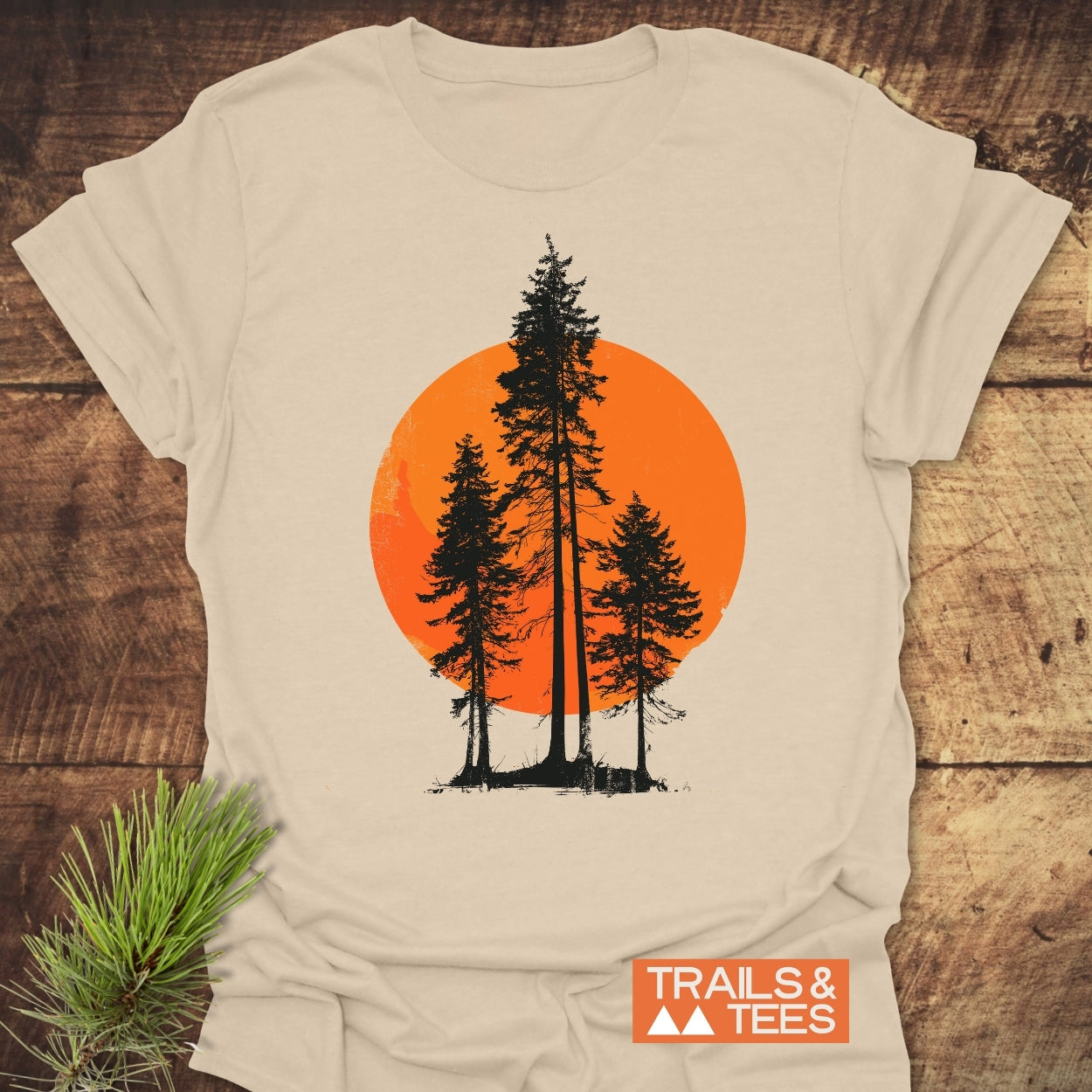 Sunset Trees T-Shirt featuring three pine silhouettes against an orange circle, laid on wood. Crafted from soft cotton, ideal for outdoor lovers. Includes “TRAILS & TEES” promotional tag.