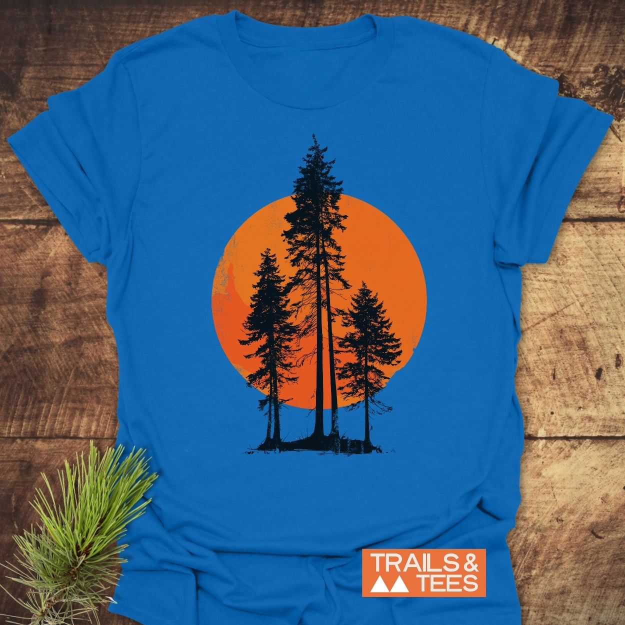 Sunset Trees T-Shirt features three pine-tree silhouettes against an orange circle, printed on bright blue cotton. Logo TRAILS & TEES visible. Laid on wood with pine needle prop.