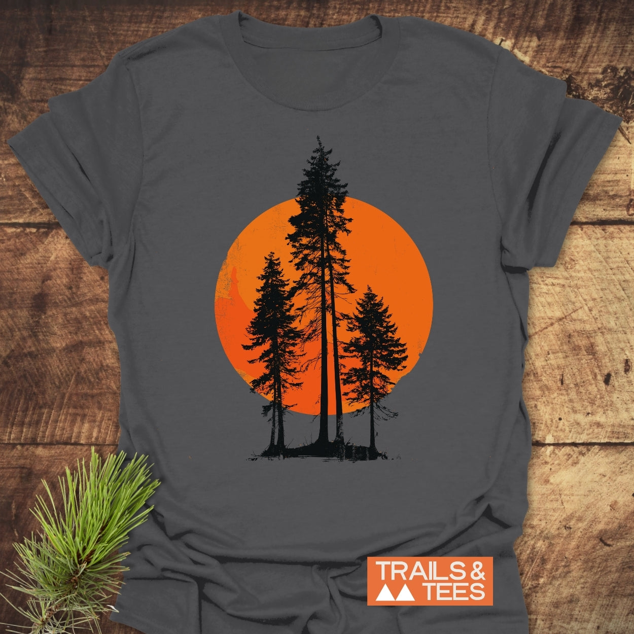 Sunset Trees T-Shirt on wooden surface featuring black pine silhouettes against an orange sun. Includes brand label TRAILS & TEES and a pine sprig, highlighting outdoor-inspired design and adventure-ready comfort.