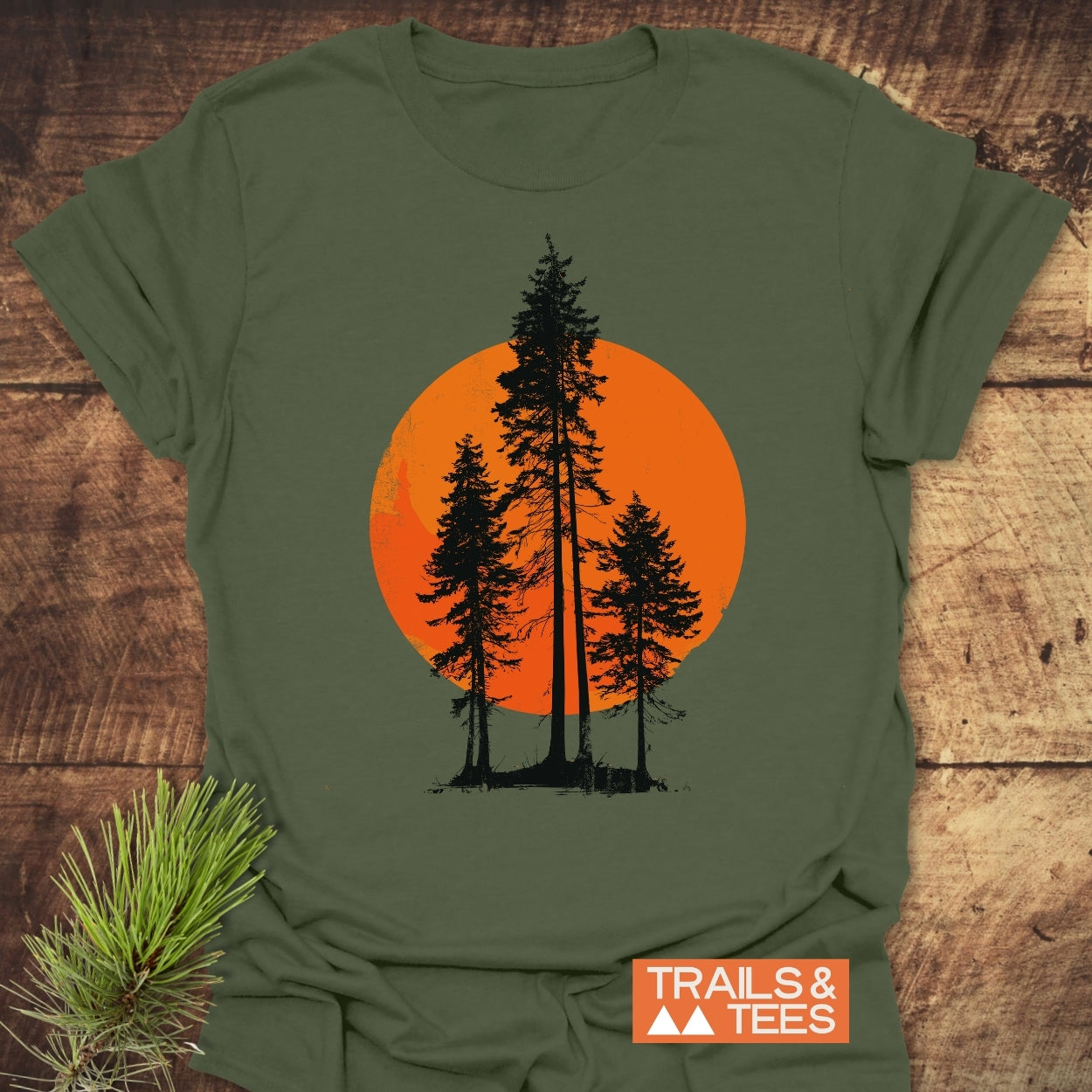 Sunset Trees T-Shirt featuring black pine-tree silhouettes against an orange circle, symbolizing a sunset or moon, on an olive-green cotton tee, laid on a rustic surface with TRAILS & TEES logo.