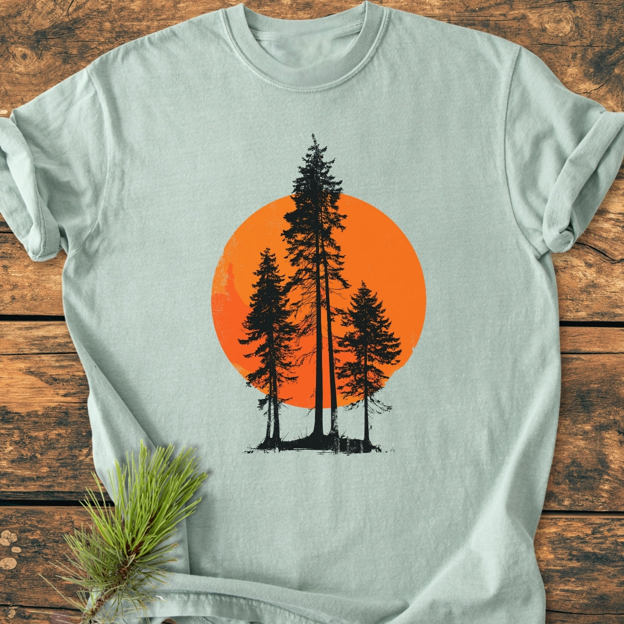 Sunset Trees - Vintage Wash Tee: Light-sage green T-shirt featuring black pine-tree silhouettes against an orange sun, displayed on wooden surface with a pine twig nearby, highlighting its cozy, relaxed fit.