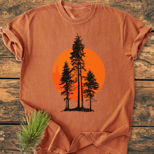 Sunset Trees - Vintage Wash Tee displayed on a wooden surface, features pine-tree silhouettes against an orange sun. Accented with a pine branch, highlighting the tee’s relaxed fit and durable, soft-washed cotton fabric.