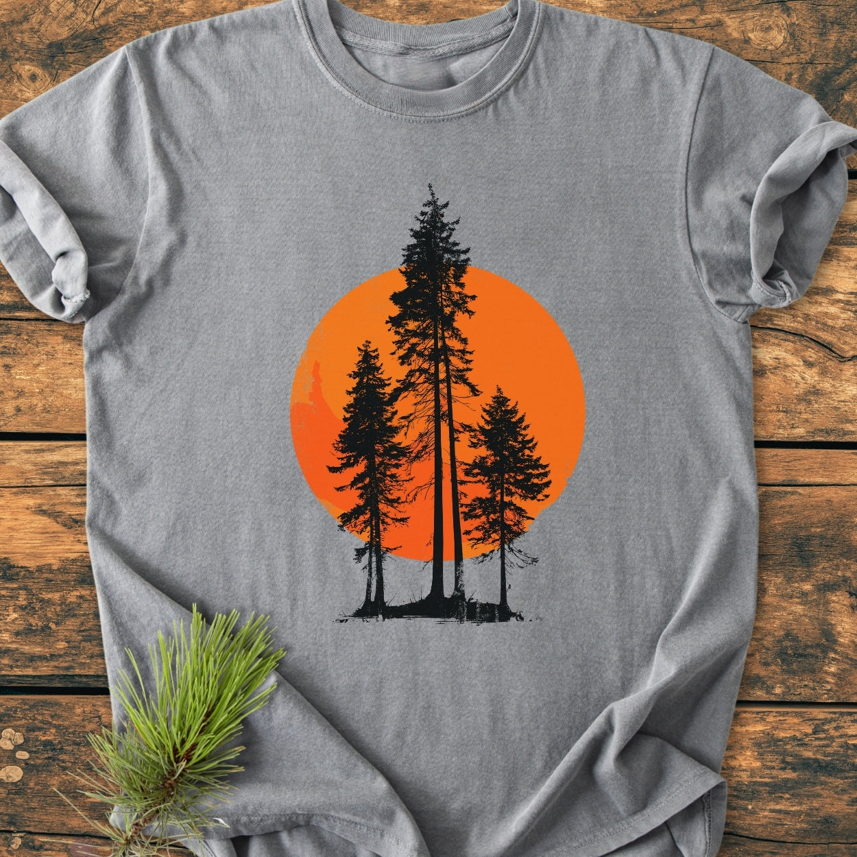 Sunset Trees - Vintage Wash Tee showcases a gray T-shirt featuring three silhouetted pine trees against an orange sun. Laid on wood, it includes a sprig of pine needles, highlighting its nature-inspired design.