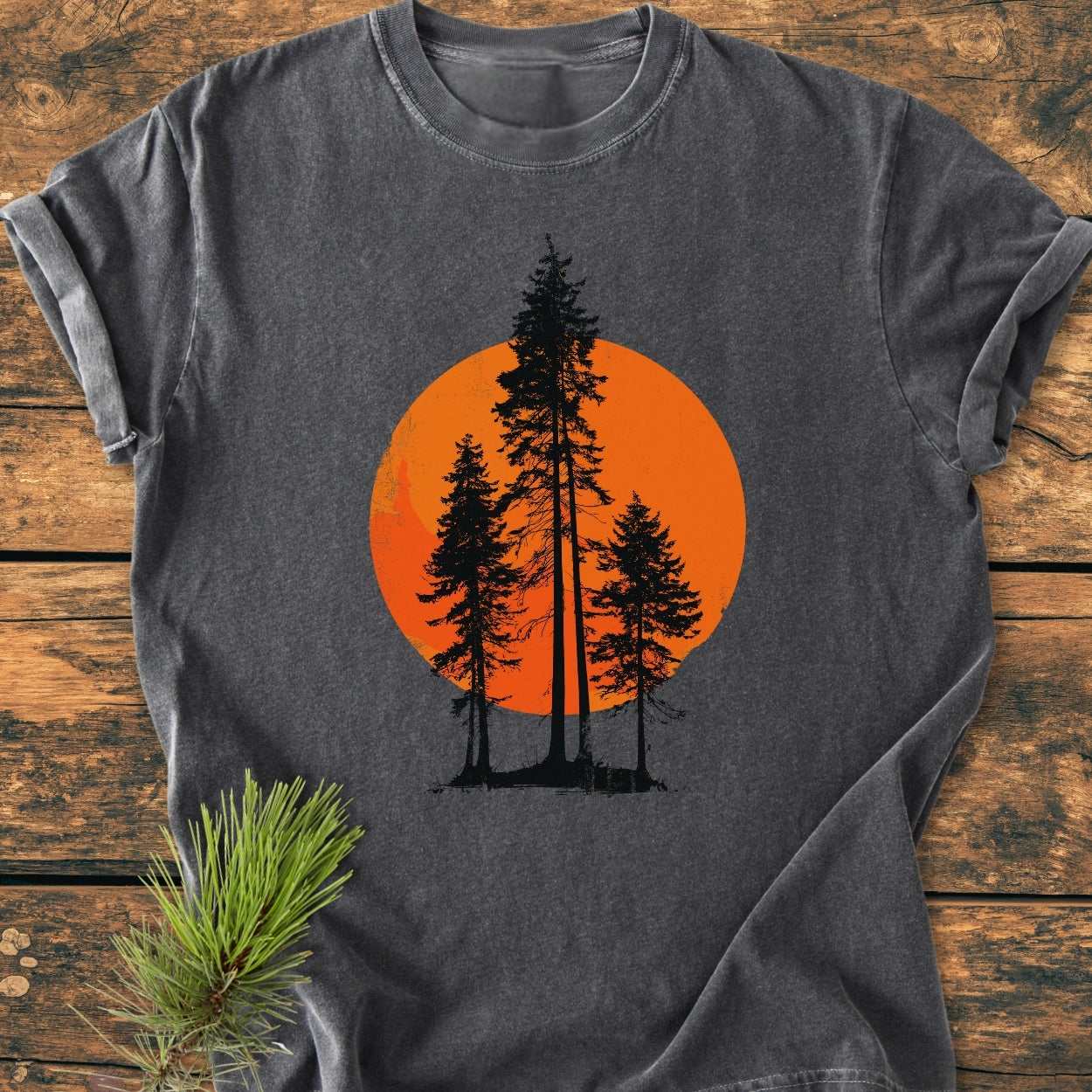 Sunset Trees - Vintage Wash Tee: Charcoal-gray T-shirt with black pine silhouettes against an orange circle, laid on wood. Features include garment-dyed, 100% ring-spun cotton, relaxed fit, and double-needle stitching.