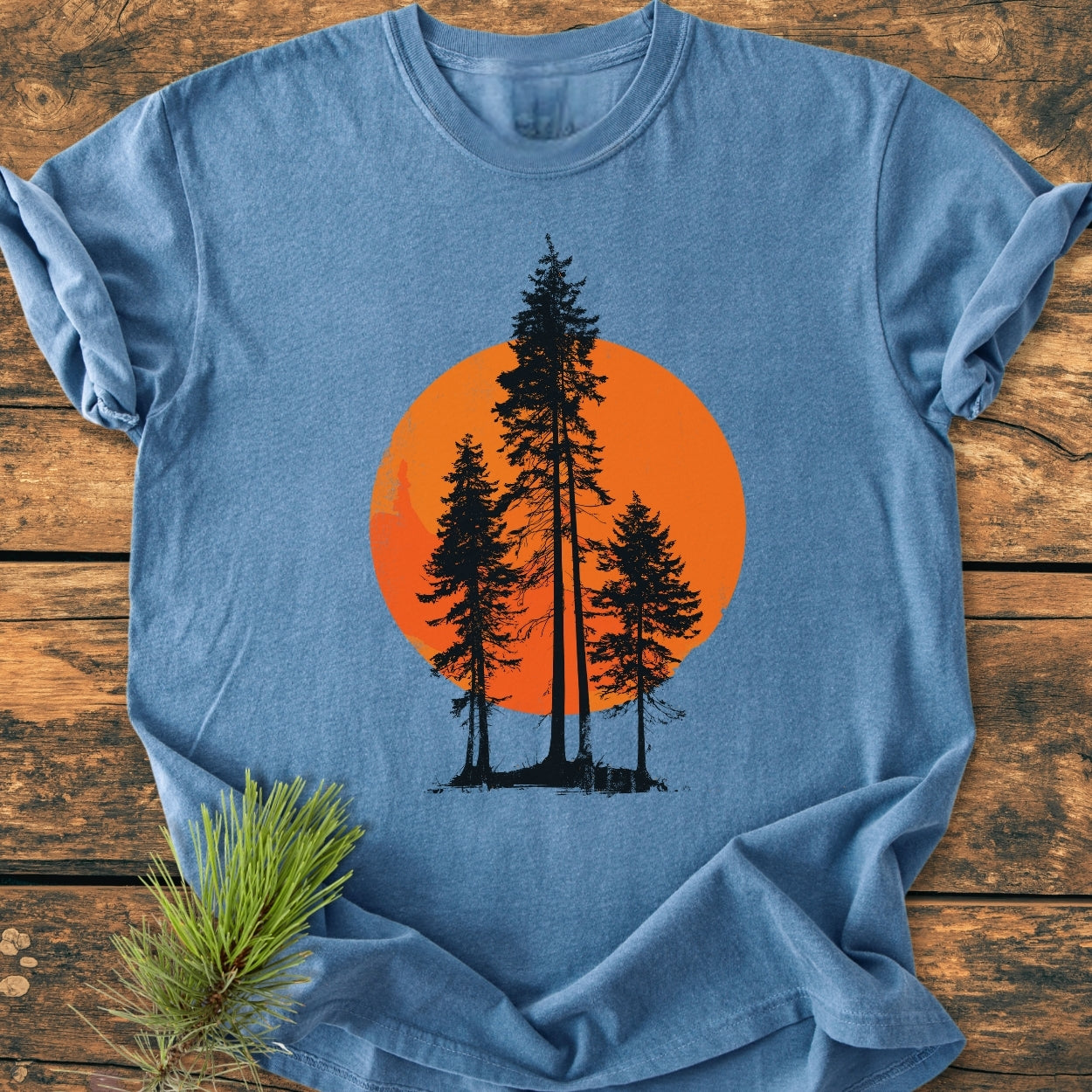 Sunset Trees - Vintage Wash Tee: Light-blue T-shirt with black pine-tree silhouettes against a large orange circle, laid out on wood. Includes a pine needle sprig accentuating the natural theme.