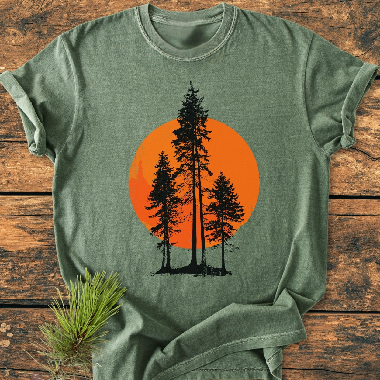 Sunset Trees - Vintage Wash Tee features four silhouetted pine trees against a large orange circle. Displayed on rustic wood, this heather-green tee offers a cozy, relaxed fit with durable double-needle stitching.