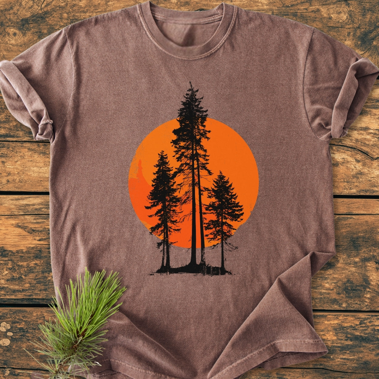 Sunset Trees - Vintage Wash Tee showcases a stylized forest scene with black pine silhouettes against an orange circle, laid flat on weathered wood, featuring a rustic pine sprig accent.