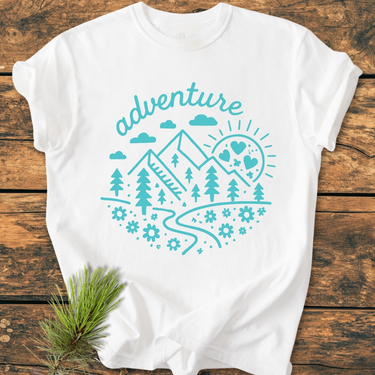 Adventure Whimsical - Vintage Wash Tee featuring a turquoise circular graphic of mountains, trees, and sun. Laid on a wooden surface, highlighting its relaxed fit and garment-dyed softness.