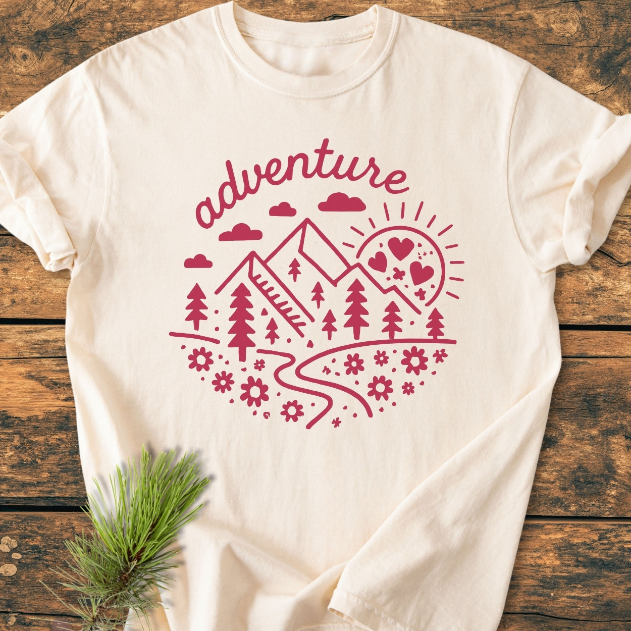 Adventure Whimsical - Vintage Wash Tee features a cream color with a dark-pink circular motif of mountains and nature elements, laid flat on wood, showcasing its soft, relaxed fit and detailed design.