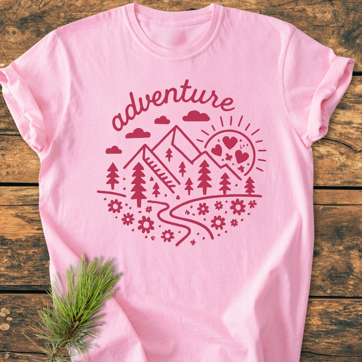 Adventure Whimsical - Vintage Wash Tee: Light pink T-shirt with a circular “adventure” graphic featuring mountains, rising sun, and daisy-like flowers, displayed on a wooden surface with a sprig of evergreen needles.