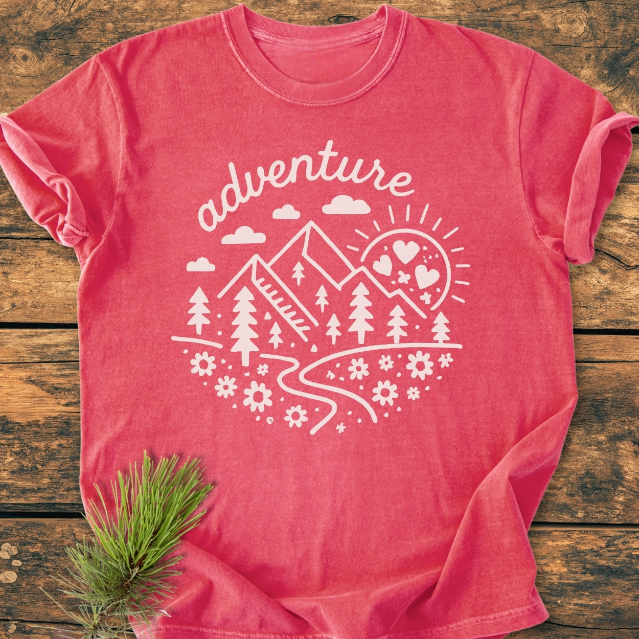 Adventure Whimsical - Vintage Wash Tee features a pink-red hue with adventure text above a nature scene, showcasing mountains, trail, trees, clouds, flowers, and heart-studded sun on soft, garment-dyed cotton.