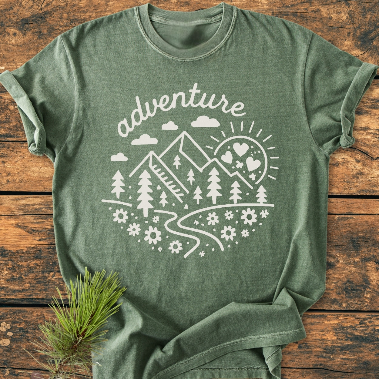 Adventure Whimsical - Vintage Wash Tee features a heather-green color with a white print of mountains, pine trees, and sun, showcasing a nature scene. Laid on wood, with pine needles beside.