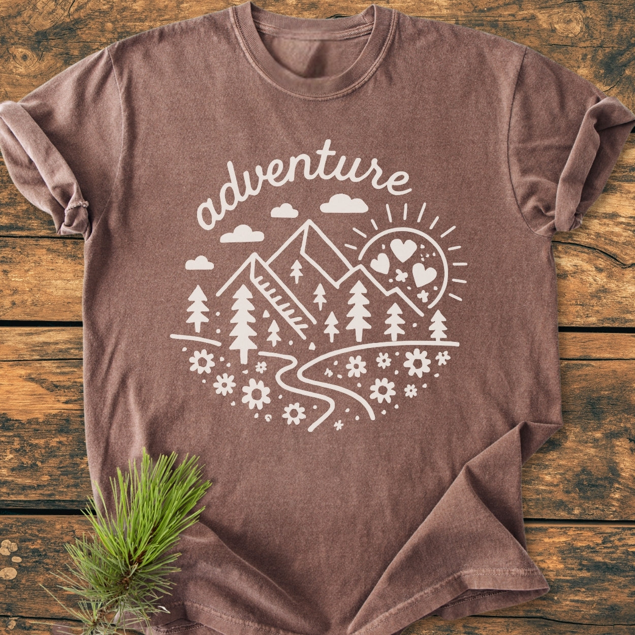Adventure Whimsical - Vintage Wash Tee features a hand-drawn scene with mountains and trees on a washed-brown crew-neck, laid flat with a decorative pine twig beside it.