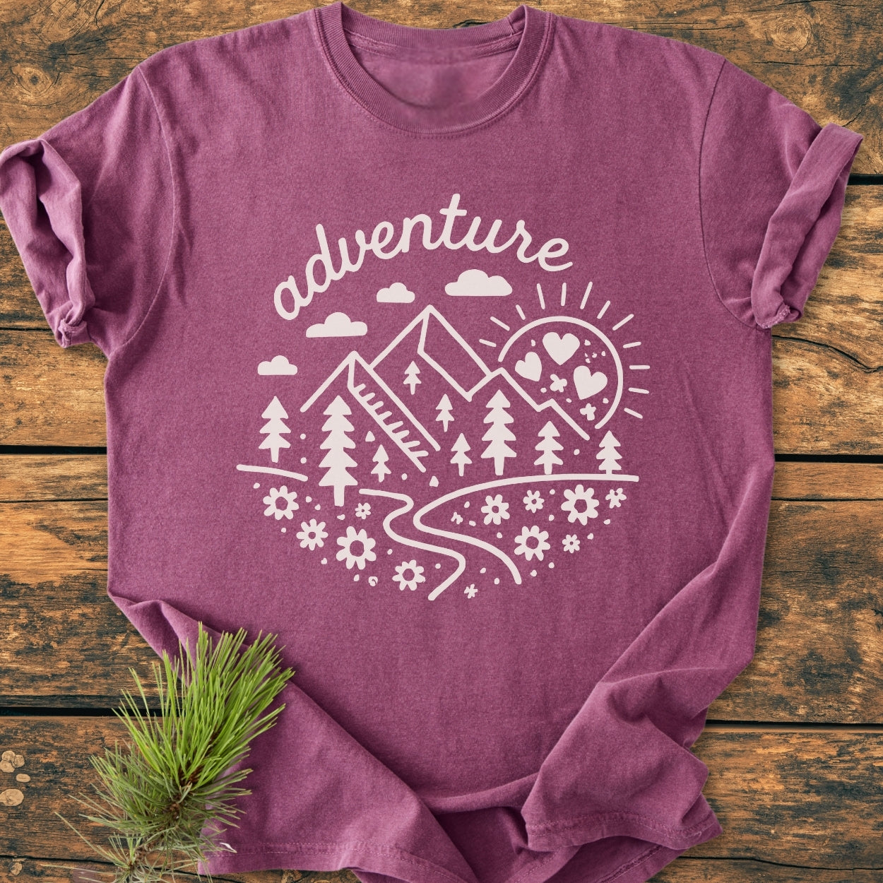 Adventure Whimsical - Vintage Wash Tee features a plum-purple hue with an off-white circular graphic of mountains, trees, and clouds. Displayed on rustic wood, it highlights style and comfort.