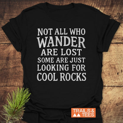 Black Not All Who Wander Are Lost - Some Are Just Looking for Cool Rocks T-shirt on wood. Features white text, orange TRAILS & TEES logo, pine sprig; embodies adventure spirit.