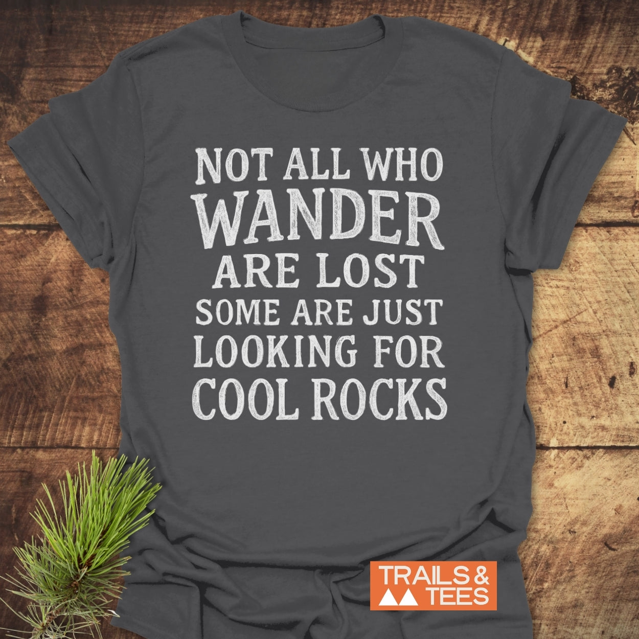 Not All... Rustic Chalkboard T-Shirt on wooden surface with pine sprig; features white distressed text. Orange TRAILS & TEES logo with mountains in corner, reflecting outdoor adventure spirit and sustainability.