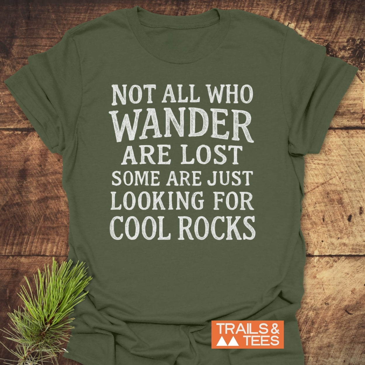 Not All... Rustic Chalkboard T-Shirt on wooden surface, featuring bold white text about wandering and rocks, with Trails & Tees logo. Beside it, a pine sprig accentuates the outdoor theme.