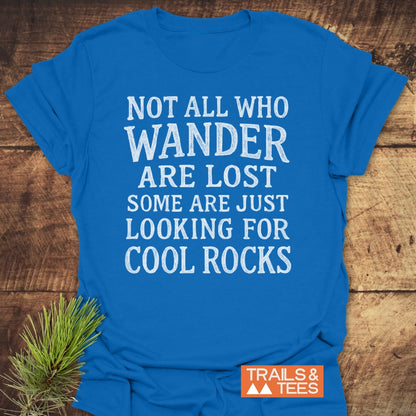 Not All... Rustic Chalkboard T-Shirt in bright blue, featuring white slogan text about wandering and rocks, with an orange TRAILS & TEES logo, laid on a wooden surface with pine sprig.