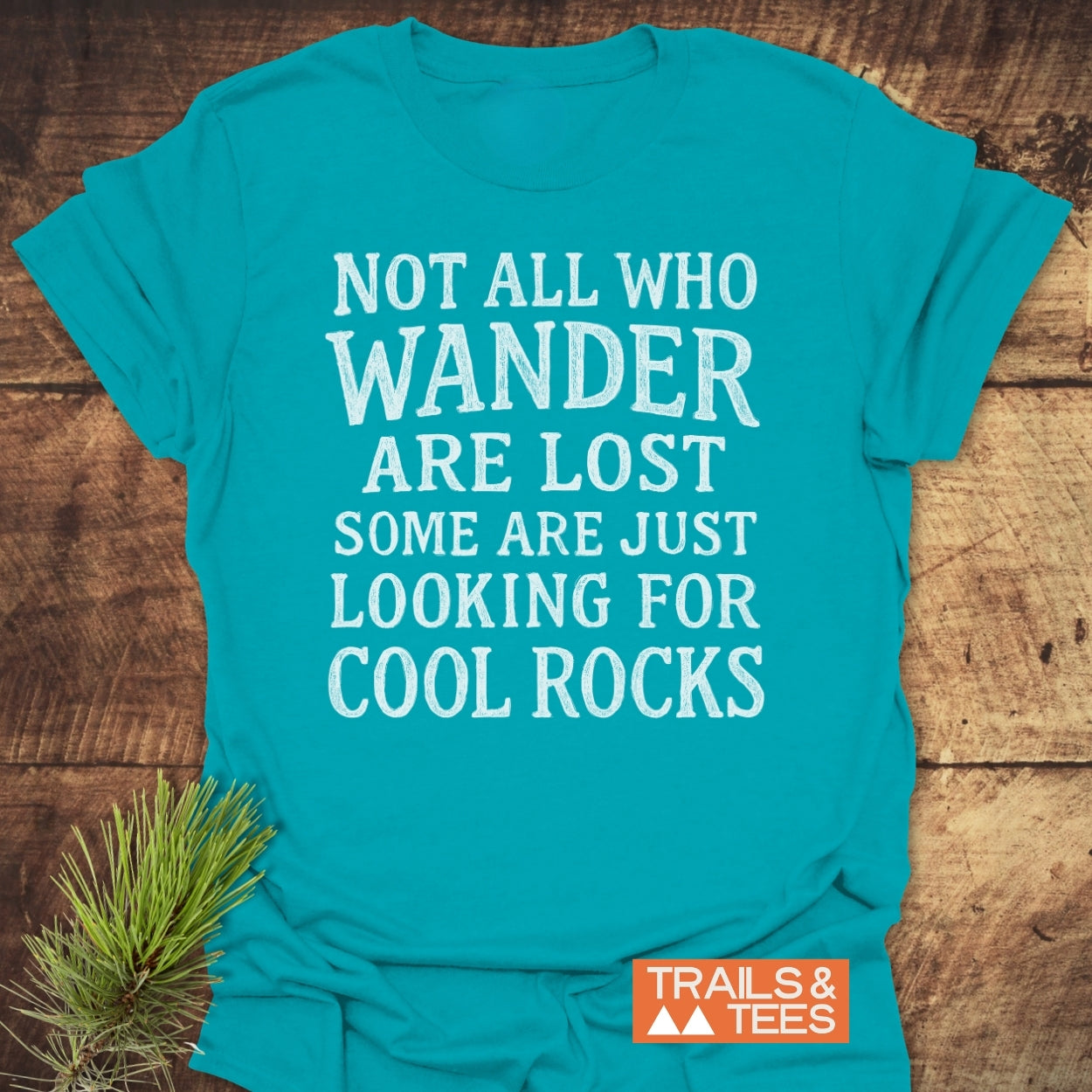 Turquoise Not All Who Wander Are Lost T-shirt in white distressed letters, on rustic wood, featuring a small orange TRAILS & TEES logo and pine needle accent beside it.