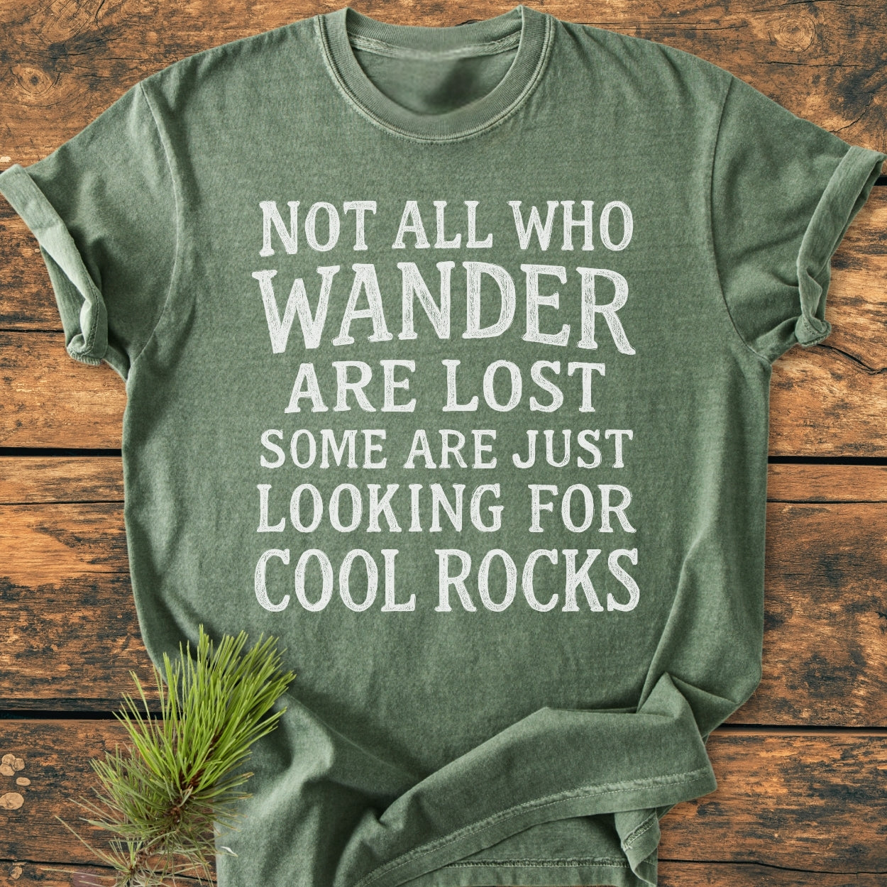 Forest-green T-shirt, Not All Who Wander Are Lost slogan in vintage font, folded on wood surface. Styled with pine branch. Not All... Rustic Chalkboard - Vintage Wash Tee emphasizes comfort and durability.